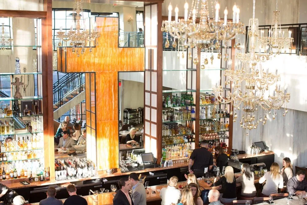 Interior of a restaurant bar with chandeliers, people seated at the bar, and a large orange decorative cross on the wall.