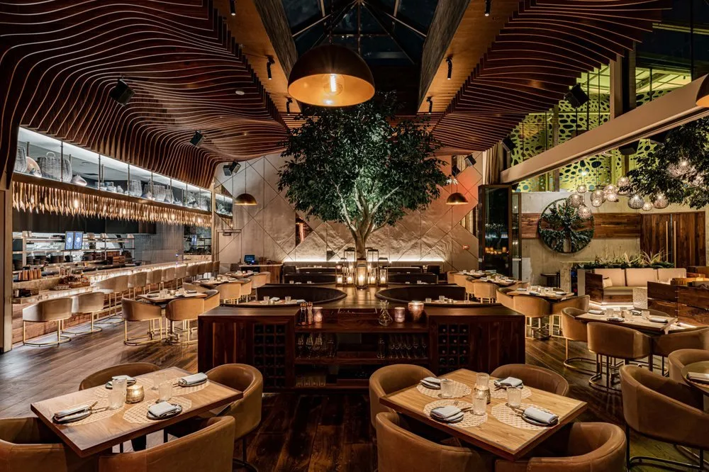 Interior of a modern, upscale restaurant with warm lighting, a large central tree, and a mix of seating options including tables with chairs and a bar area.