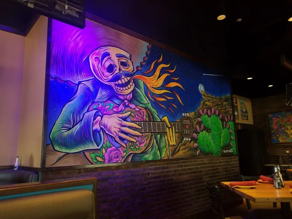 Colorful mural of a skeleton playing guitar with flames coming from its mouth, set against a starry night sky, mountains, and cacti.