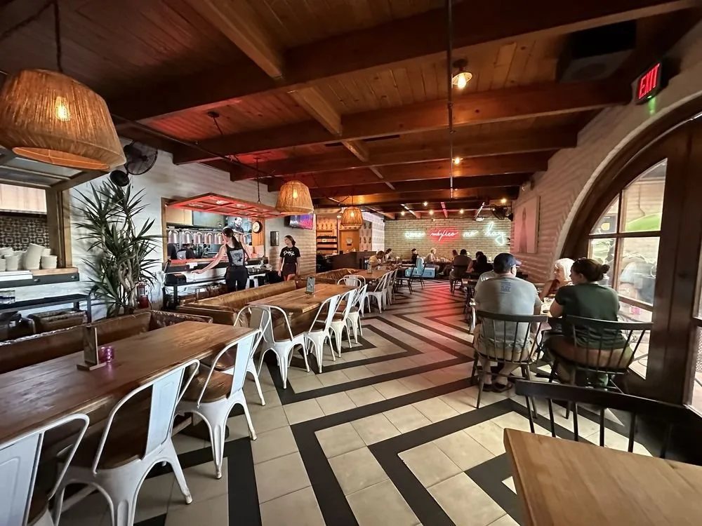 Inside a modern restaurant with wooden ceilings and stylish lighting, several tables are occupied by customers, and a bar area with staff serving drinks is visible in the background.
