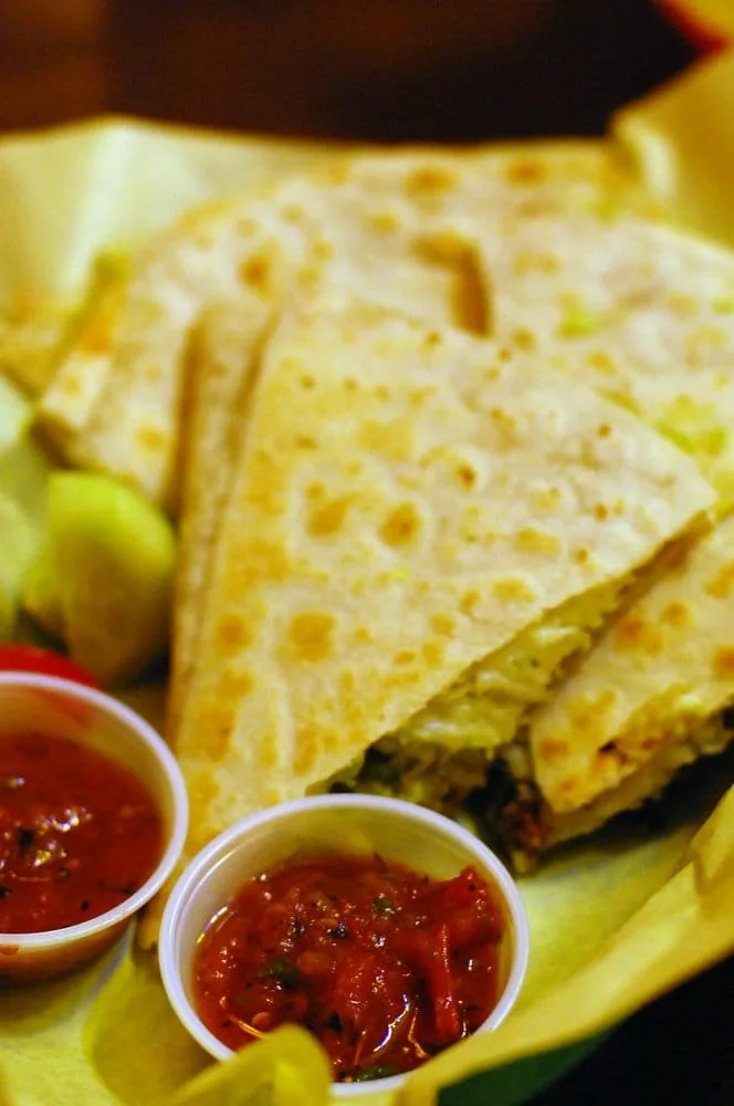 Close-up of a large quesadilla cut into sections, served with two cups of salsa and lemon wedges on the side.