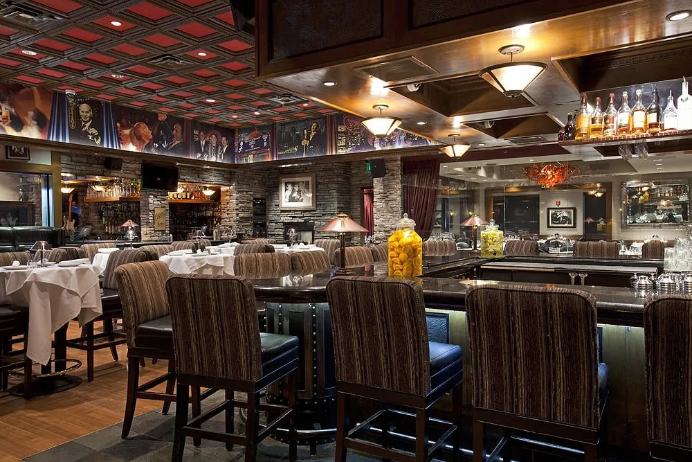 Interior of an upscale restaurant with tables covered in white tablecloths, a bar with chairs, a brick accent wall, and decorative jars filled with lemons and limes.