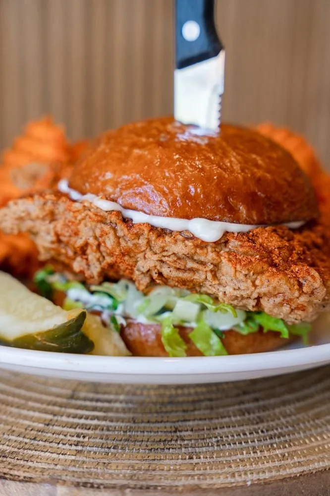 Close-up of a crispy fried chicken sandwich with mayonnaise, lettuce, and pickles on a bun, with a knife inserted into the top bun.