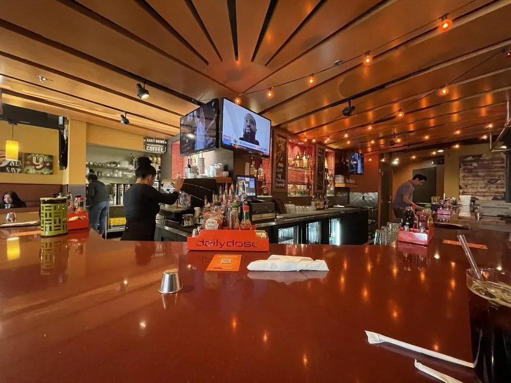 Interior of a cozy bar or cafe with a wooden ceiling, dim warm lighting, a bartender preparing drinks, and a customer working behind the bar. There are multiple TV screens on the walls and various bottles and bar equipment visible.