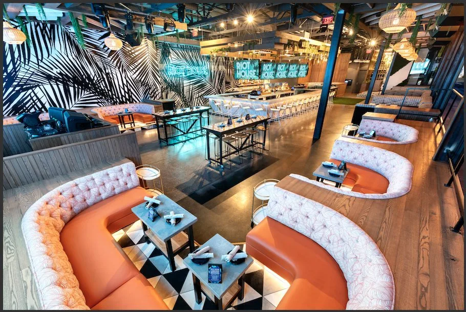 Interior of a modern restaurant or bar with curved seating booths, a long bar counter with barstools, decorative lighting, and a large digital sign saying 'Riot House' in the background.