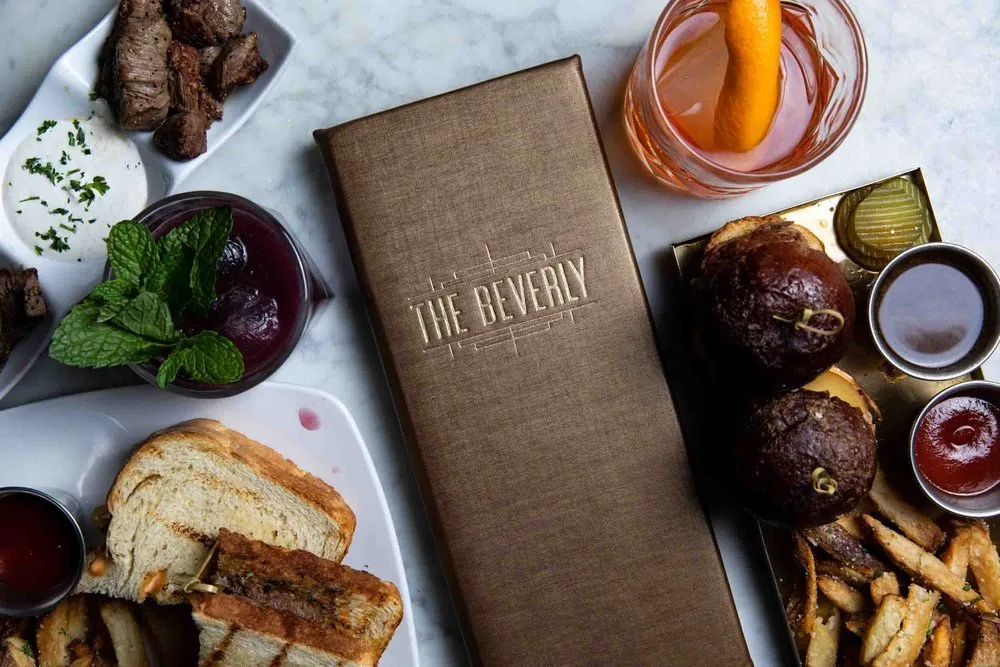 A meal spread includes a sandwich, some steaks, fries, dipping sauces, a drink with a lemon twist, a bowl of cherries with mint, and a tray of sliders with condiments, all on a marble surface.